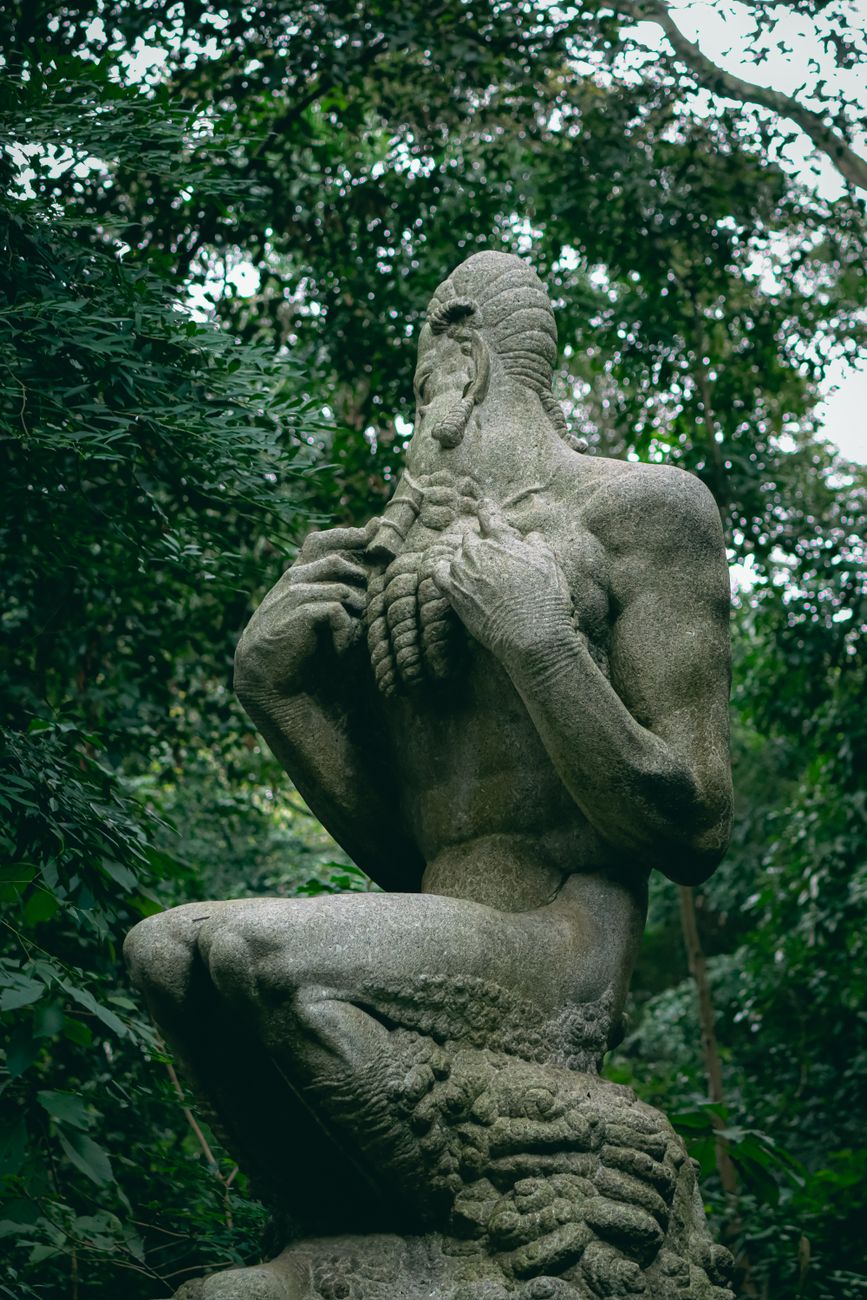 A stone statue of a humanoid figure with aquatic features, kneeling amidst greenery. The figure has textured skin, a muscular build, and appears to be holding something in its hands.